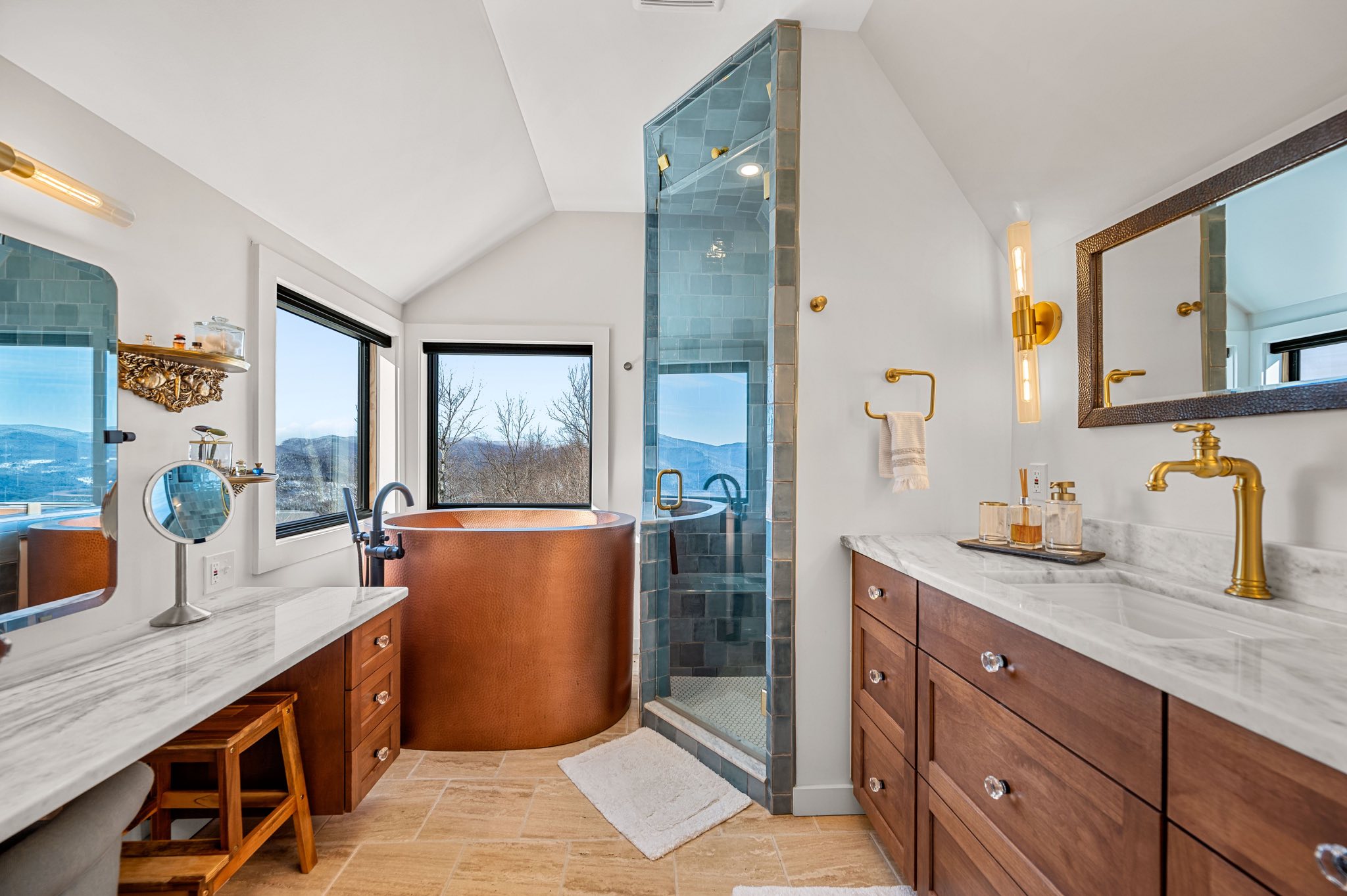 Hammered copper soaking tub in a bathroom remodel with marble vanities, blue tiled shower, and mountain windows